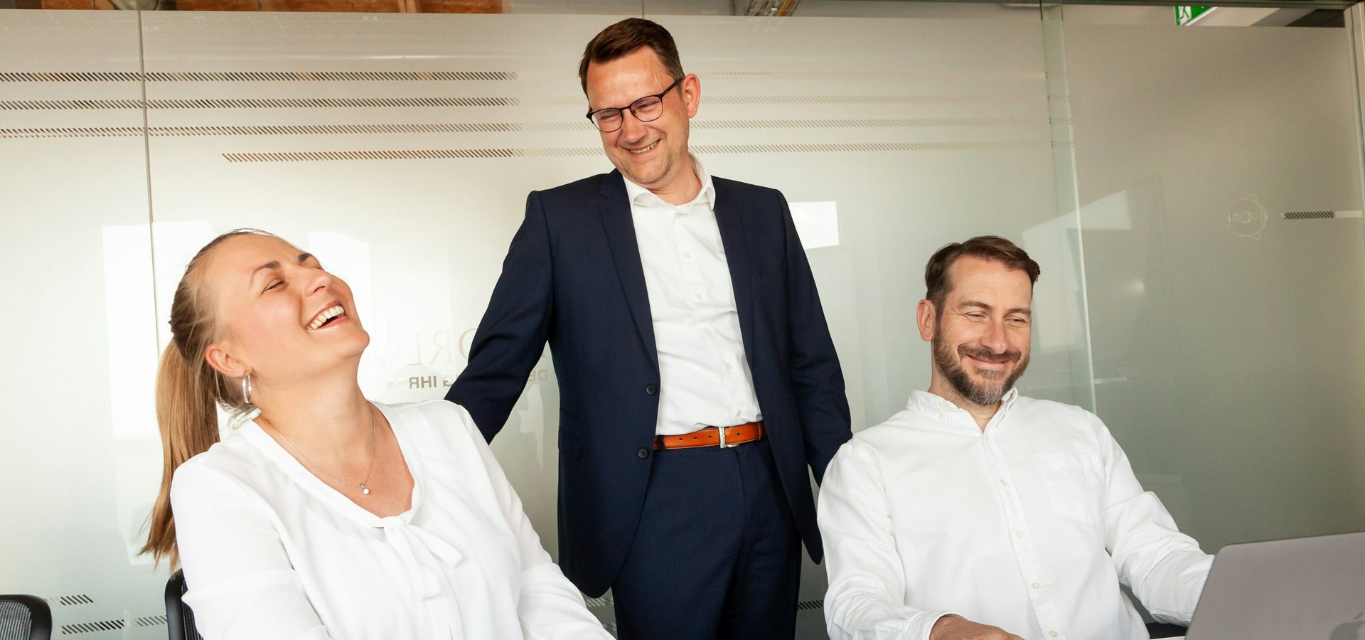 Three colleagues laughing at a laptop in a glass-walled office