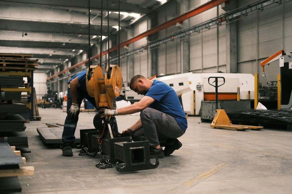 Two technicians attach metal parts to the hook of an overhead crane in a workshop