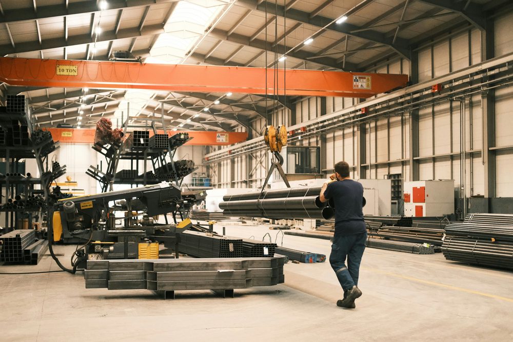 An operator guides steel sections lifted by an overhead crane in an industrial hall